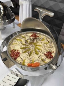 Creamy hummus served in a stainless steel chafing dish, garnished with olive oil drizzle, black pepper, pomegranate seeds, and carrot slices at a buffet setup.