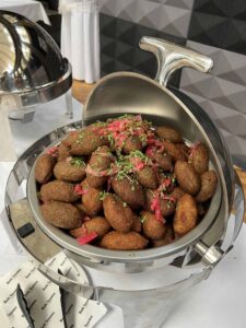 Golden-brown fried kibbeh arranged in a stainless steel chafing dish, garnished with chopped herbs and pickled red onions at a buffet.