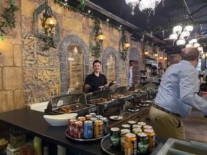 Traditional Middle Eastern restaurant buffet with hot food trays, colorful drinks, decorative stone walls, and warm hanging lanterns.